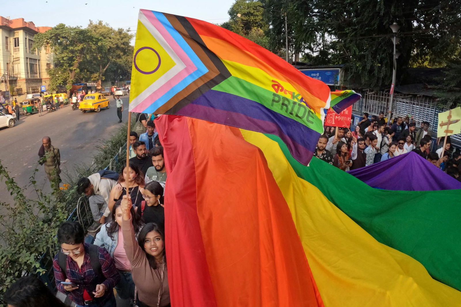 Crowds gather on a city street for an LGBTQ+ pride march, holding up a large rainbow flag alongside a Progress Pride flag that reads 'Pride' in English and an Asian script. The scene celebrates diversity and inclusivity during Asian Pride Month, with participants of various backgrounds coming together in support.